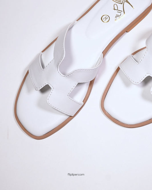 White slip-on sandals with gold detailing placed on a wooden table beside the ocean