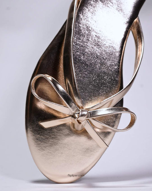 Person wearing gold bow-detail sandals while standing on a beige floor