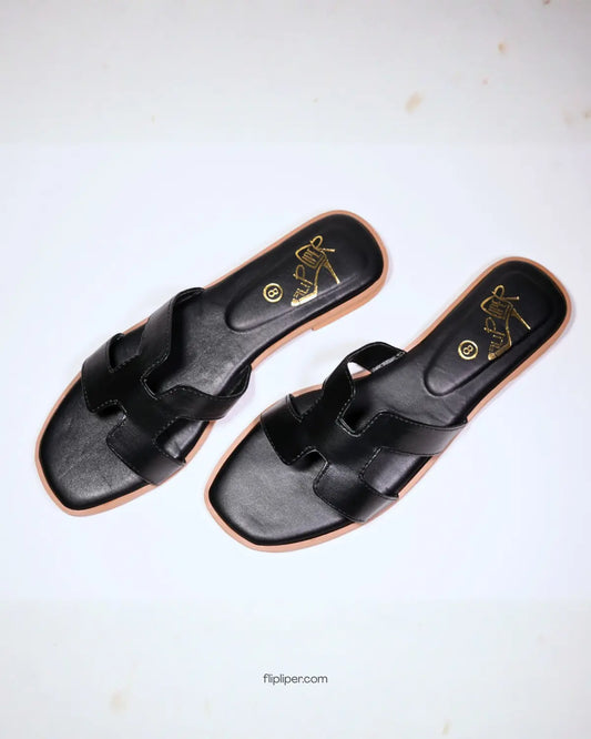 Black slip-on sandals with tan wooden soles displayed on a light beige background with minimal props