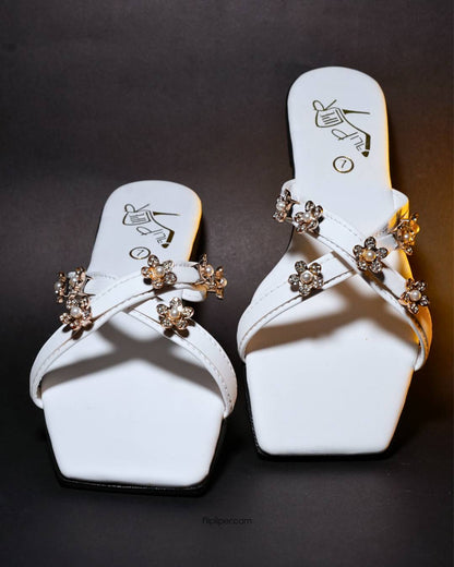 White sandals with gold floral embellishments displayed on a beige fabric background
