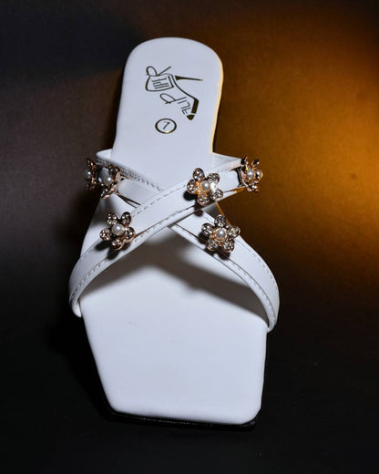 White sandals with gold floral embellishments displayed on a beige fabric background
