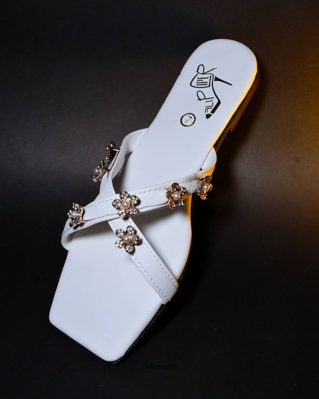 White sandals with gold floral embellishments displayed on a beige fabric background
