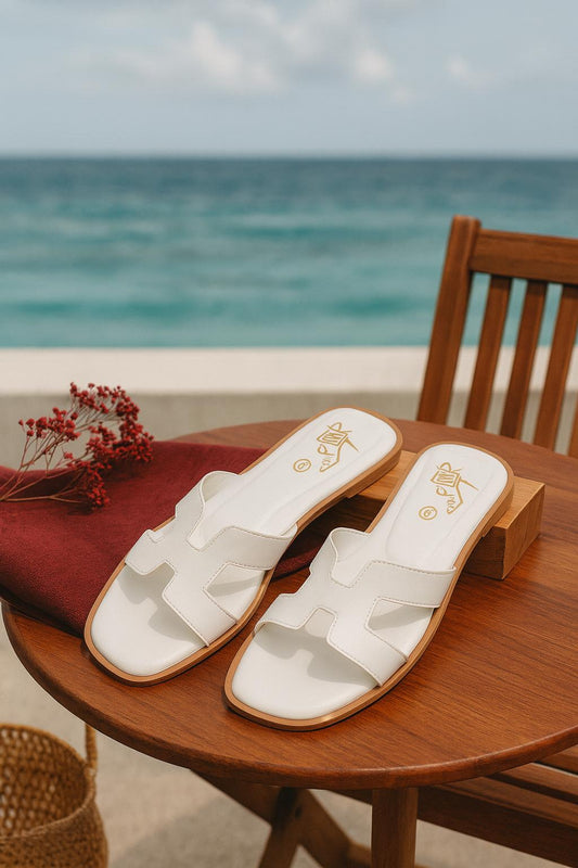 White slip-on sandals with gold detailing placed on a wooden table beside the ocean
