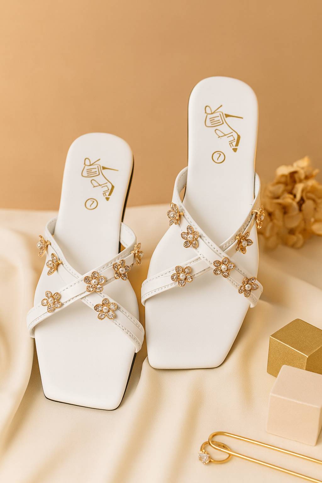White sandals with gold floral embellishments displayed on a beige fabric background
